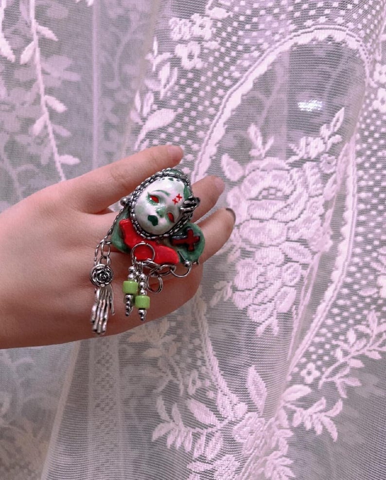 May include: A unique ring featuring a doll face with red eyes and a green face. The ring has a red bow, green beads, and a silver hand charm. The ring is held up against a white lace curtain.