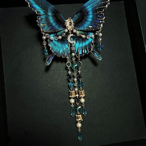 Puede incluir: Un broche decorativo de mariposa con alas azul iridiscente y bordes negros. El centro est&aacute; adornado con un peque&ntilde;o dije de media luna, cristales transparentes y perlas blancas. Tres cadenas colgantes presentan cuentas de cristal azul y detalles dorados.