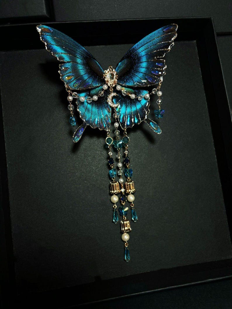 Puede incluir: Un broche decorativo de mariposa con alas azul iridiscente y bordes negros. El centro est&aacute; adornado con un peque&ntilde;o dije de media luna, cristales transparentes y perlas blancas. Tres cadenas colgantes presentan cuentas de cristal azul y detalles dorados.