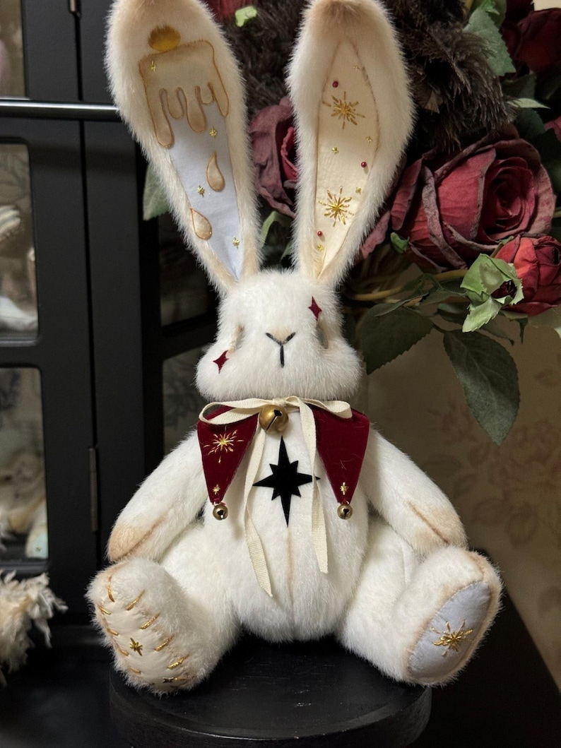 Puede incluir: Un conejo de peluche blanco con orejas largas y decoradas. Las orejas presentan detalles dorados y un dise&ntilde;o de goteo en una oreja y estrellas en la otra. Lleva un collar rojo y negro con una campana y una estrella. Las patas del conejo est&aacute;n bordadas con estrellas doradas.