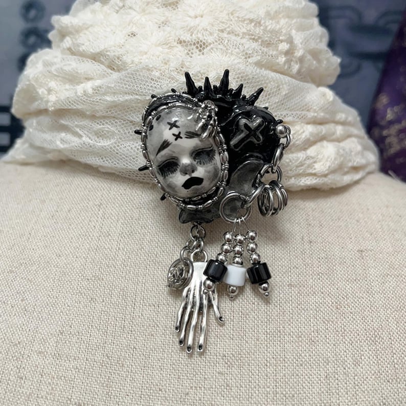 May include: A gothic-style brooch featuring a doll face, a heart with a cross, and dangling charms. The brooch is primarily black and silver, with accents of white and black beads. The doll face has stitched eyes and a sad expression.