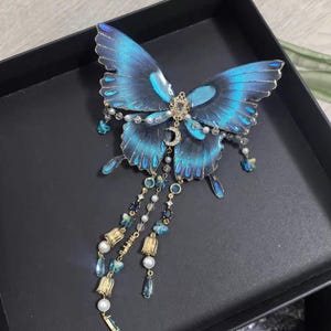 Puede incluir: Un accesorio decorativo para el cabello con forma de mariposa, con alas de color azul vibrante y bordes dorados. El centro est&aacute; adornado con una peque&ntilde;a luna creciente dorada y cristales transparentes. Tres cadenas colgantes incluyen perlas blancas, cristales azules y dijes dorados en forma de campana.