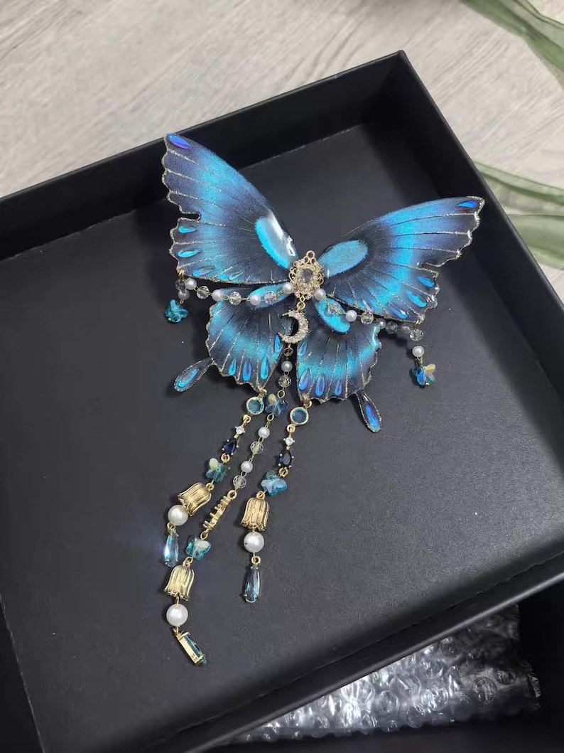 Puede incluir: Un accesorio decorativo para el cabello con forma de mariposa, con alas de color azul vibrante y bordes dorados. El centro est&aacute; adornado con una peque&ntilde;a luna creciente dorada y cristales transparentes. Tres cadenas colgantes incluyen perlas blancas, cristales azules y dijes dorados en forma de campana.