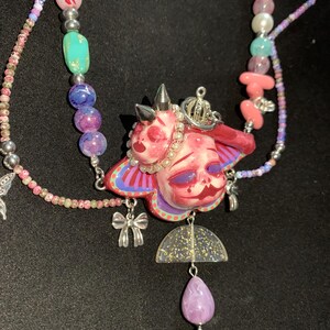 May include: A handmade necklace featuring a pink and red pendant with a face design, adorned with a silver crown and spikes. The necklace includes a variety of colorful beads, a clear half-circle pendant with gold glitter, and a purple teardrop-shaped bead.
