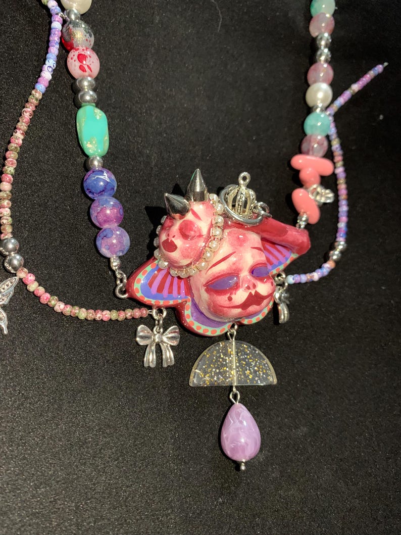 May include: A handmade necklace featuring a pink and red pendant with a face design, adorned with a silver crown and spikes. The necklace includes a variety of colorful beads, a clear half-circle pendant with gold glitter, and a purple teardrop-shaped bead.
