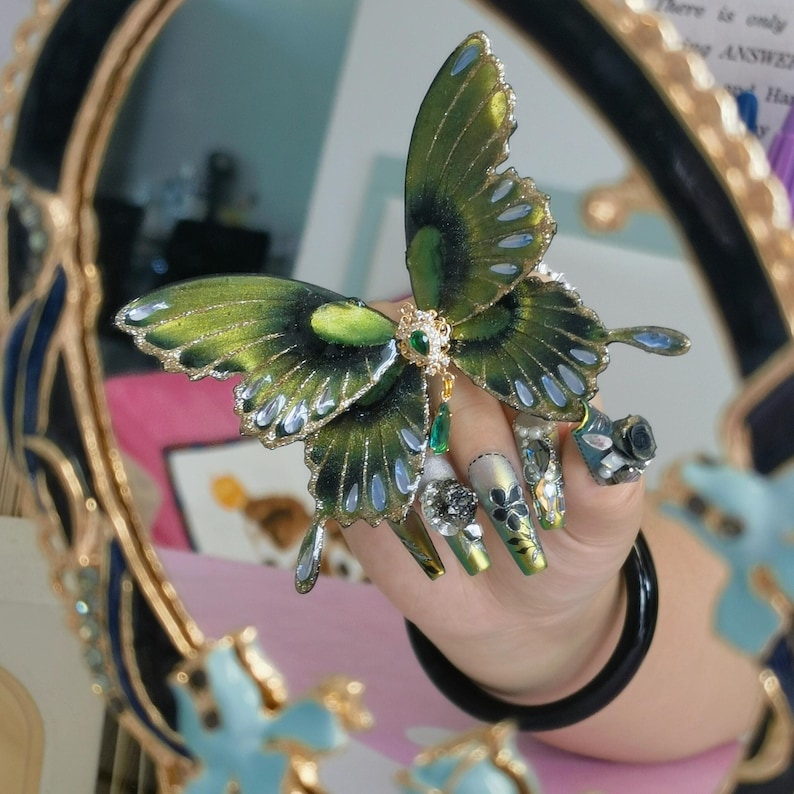 Puede incluir: Un gran adorno decorativo de mariposa con alas de color verde iridiscente y negro con bordes plateados. La mariposa est&aacute; adornada con una pieza central de filigrana dorada, una peque&ntilde;a piedra preciosa verde y un dije de gota verde.