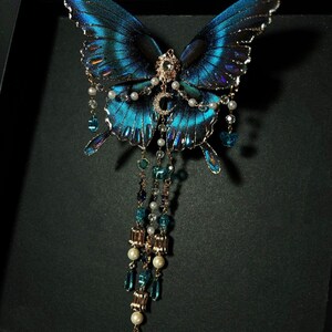 Puede incluir: Un broche decorativo de mariposa con alas de color azul iridiscente y bordes negros. El centro est&aacute; adornado con una luna creciente dorada y un peque&ntilde;o cristal transparente. Tres cadenas colgantes incluyen perlas blancas y cuentas de cristal azul.