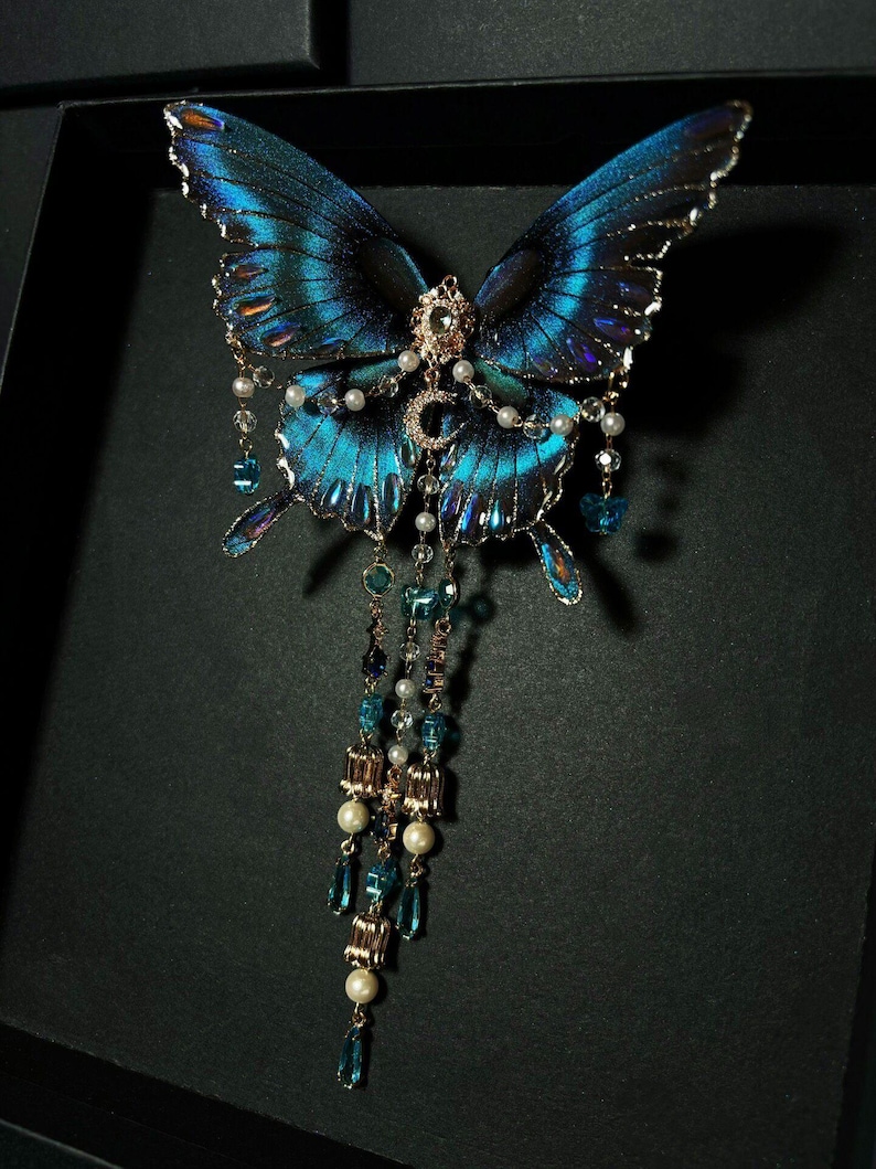 Puede incluir: Un broche decorativo de mariposa con alas de color azul iridiscente y bordes negros. El centro est&aacute; adornado con una luna creciente dorada y un peque&ntilde;o cristal transparente. Tres cadenas colgantes incluyen perlas blancas y cuentas de cristal azul.