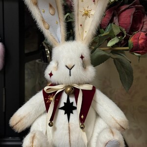 Puede incluir: Una mu&ntilde;eca de peluche de conejo blanco con orejas largas. Las orejas tienen detalles en azul claro y crema con adornos dorados. El conejo lleva un cuello burdeos y crema con una estrella negra y detalles dorados. Peque&ntilde;as campanas cuelgan del cuello.