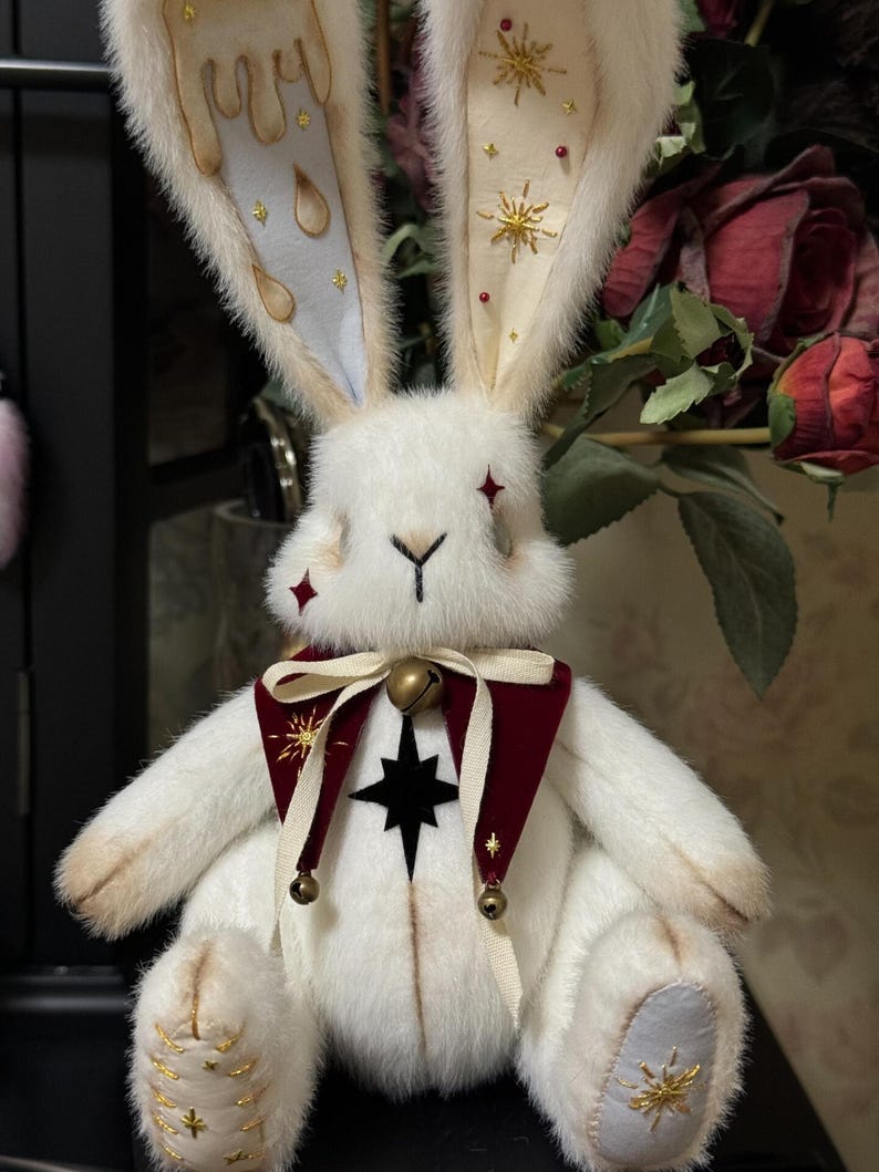 Puede incluir: Una mu&ntilde;eca de peluche de conejo blanco con orejas largas. Las orejas tienen detalles en azul claro y crema con adornos dorados. El conejo lleva un cuello burdeos y crema con una estrella negra y detalles dorados. Peque&ntilde;as campanas cuelgan del cuello.