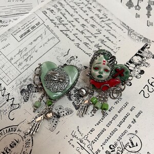 May include: Two unique, handmade brooches. One is heart-shaped, light green with a silver filigree design and a dangling silver hand. The other features a doll face with red accents and a dangling silver hand. Both have green beads.