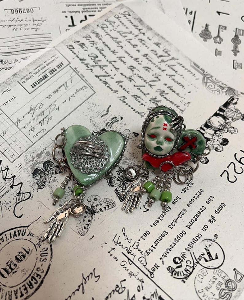 May include: Two unique, handmade brooches. One is heart-shaped, light green with a silver filigree design and a dangling silver hand. The other features a doll face with red accents and a dangling silver hand. Both have green beads.