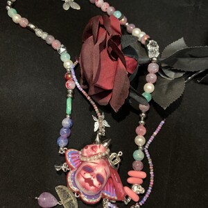 May include: A unique necklace featuring a central pendant with a skull design, adorned with a crown and butterfly accents. The necklace incorporates a variety of beads in shades of pink, purple, and green, with a dark red rose and a silver chain.