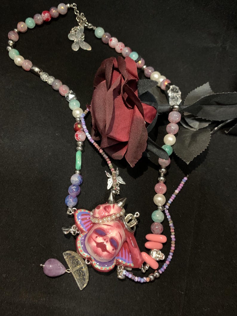 May include: A unique necklace featuring a central pendant with a skull design, adorned with a crown and butterfly accents. The necklace incorporates a variety of beads in shades of pink, purple, and green, with a dark red rose and a silver chain.