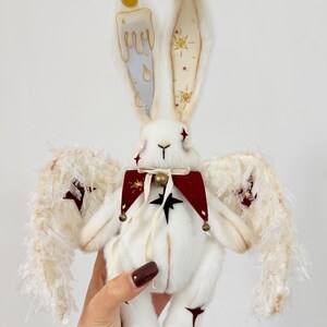 Puede incluir: Una mu&ntilde;eca de peluche de conejo blanco con orejas largas, adornada con un dise&ntilde;o de vela en una oreja y detalles de estrellas en la otra. Tiene alas de plumas, un cuello rojo y negro y una campana. La mu&ntilde;eca es sostenida por una mano.