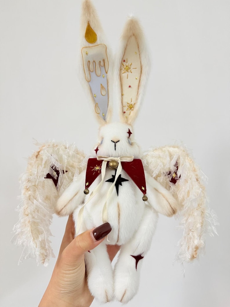 Puede incluir: Una mu&ntilde;eca de peluche de conejo blanco con orejas largas, adornada con un dise&ntilde;o de vela en una oreja y detalles de estrellas en la otra. Tiene alas de plumas, un cuello rojo y negro y una campana. La mu&ntilde;eca es sostenida por una mano.