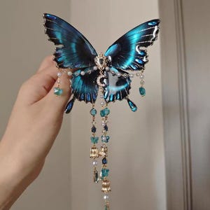 Puede incluir: Un accesorio decorativo para el cabello con una gran mariposa azul iridiscente de bordes negros. La mariposa est&aacute; adornada con un dije central de media luna, peque&ntilde;as perlas blancas y cadenas doradas colgantes con cuentas azules y dijes.