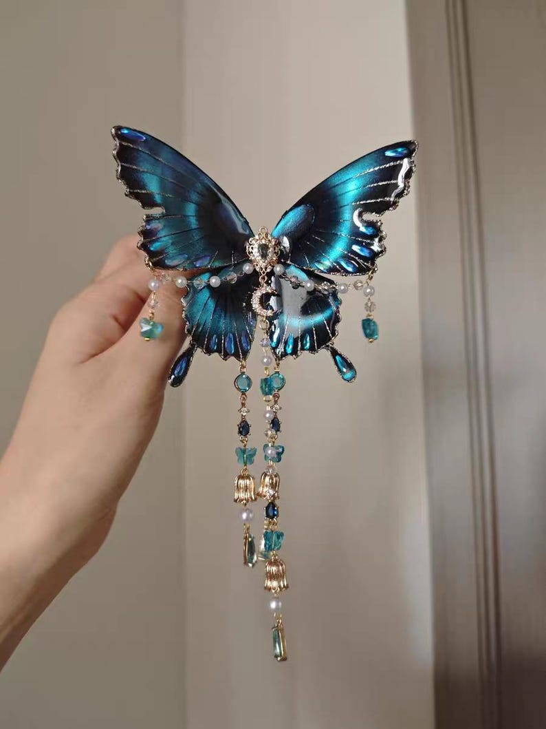 Puede incluir: Un accesorio decorativo para el cabello con una gran mariposa azul iridiscente de bordes negros. La mariposa est&aacute; adornada con un dije central de media luna, peque&ntilde;as perlas blancas y cadenas doradas colgantes con cuentas azules y dijes.