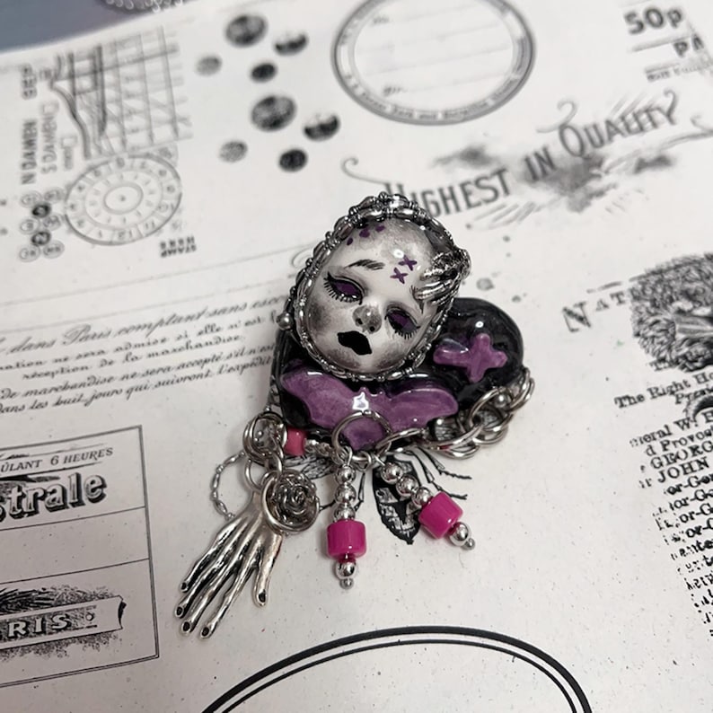 May include: A handcrafted brooch featuring a doll face with a black and purple color scheme. The brooch has a silver-toned metal frame and accents, with dangling pink beads and a hand charm. The design is intricate and detailed.