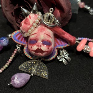 May include: A unique necklace featuring a doll-like pendant with a pink face, adorned with a crown and silver spikes. The pendant is surrounded by a colorful, floral-shaped design and includes a purple teardrop-shaped bead and a glittery umbrella charm.
