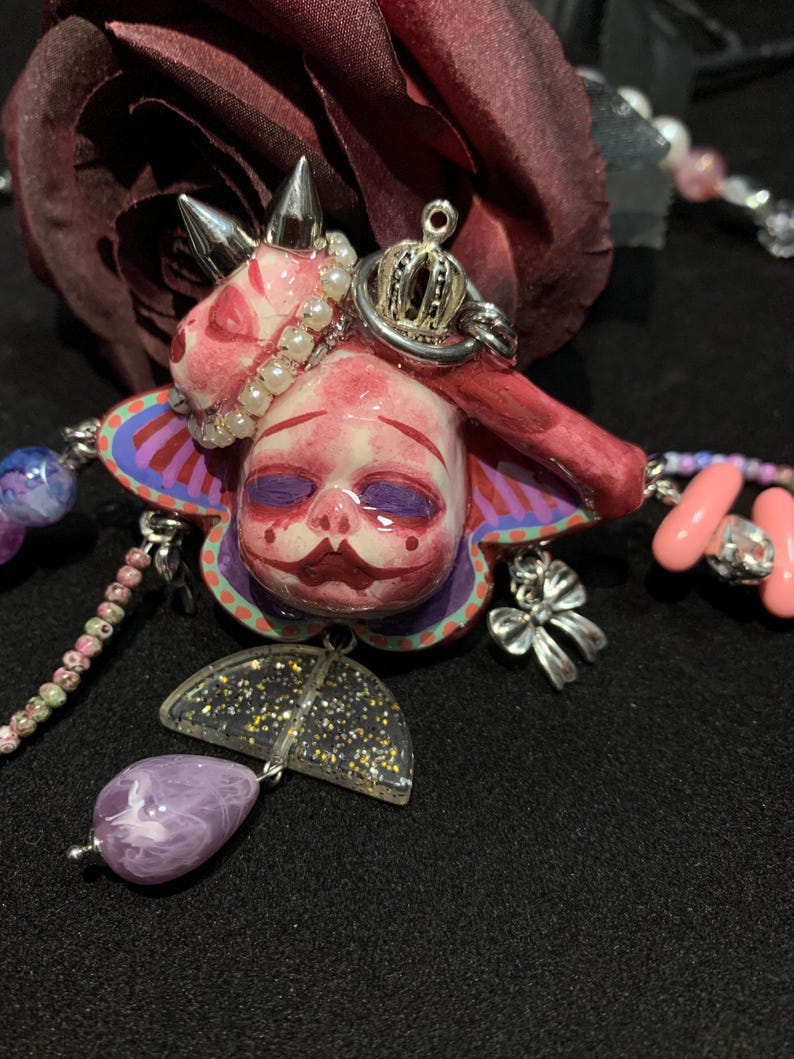 May include: A unique necklace featuring a doll-like pendant with a pink face, adorned with a crown and silver spikes. The pendant is surrounded by a colorful, floral-shaped design and includes a purple teardrop-shaped bead and a glittery umbrella charm.