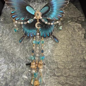 Puede incluir: Un accesorio decorativo para el cabello con forma de mariposa, con alas iridiscentes azules y negras con bordes dorados. El centro est&aacute; adornado con un dije de luna creciente dorada, cristales transparentes y perlas blancas. Tres cadenas colgantes presentan peque&ntilde;os dijes de mariposas azules, detalles met&aacute;licos dorados y perlas blancas.