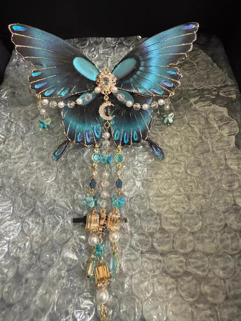 Puede incluir: Un accesorio decorativo para el cabello con forma de mariposa, con alas iridiscentes azules y negras con bordes dorados. El centro est&aacute; adornado con un dije de luna creciente dorada, cristales transparentes y perlas blancas. Tres cadenas colgantes presentan peque&ntilde;os dijes de mariposas azules, detalles met&aacute;licos dorados y perlas blancas.