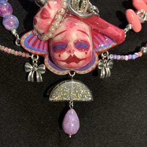 May include: A unique necklace featuring a pink and red face pendant with a silver crown and bow charms. The necklace includes a purple teardrop-shaped bead and a half-circle pendant with gold glitter. The necklace is strung with various colorful beads.
