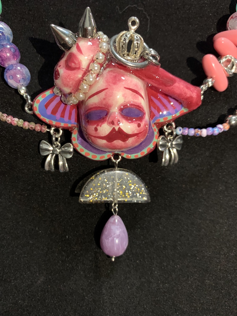 May include: A unique necklace featuring a pink and red face pendant with a silver crown and bow charms. The necklace includes a purple teardrop-shaped bead and a half-circle pendant with gold glitter. The necklace is strung with various colorful beads.