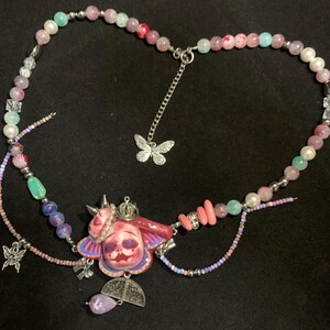 May include: A handcrafted necklace featuring a central pendant with a face design, adorned with a crown and butterfly accents. The necklace incorporates a variety of beads in shades of pink, blue, and white, with silver-toned chain details.