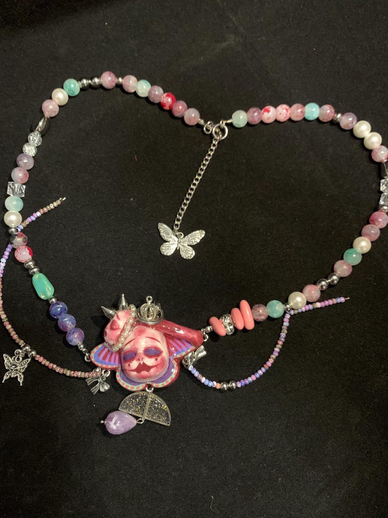 May include: A handcrafted necklace featuring a central pendant with a face design, adorned with a crown and butterfly accents. The necklace incorporates a variety of beads in shades of pink, blue, and white, with silver-toned chain details.