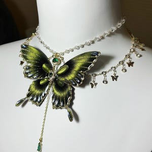 Puede incluir: Un collar de varias capas con un gran colgante de mariposa de tela verde oscuro con detalles met&aacute;licos y una piedra verde central. El collar incluye una cadena delicada con peque&ntilde;as cuentas transparentes y dijes de mariposa en tono dorado.