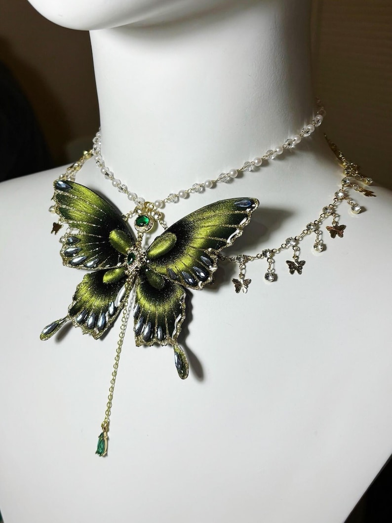 Puede incluir: Un collar de varias capas con un gran colgante de mariposa de tela verde oscuro con detalles met&aacute;licos y una piedra verde central. El collar incluye una cadena delicada con peque&ntilde;as cuentas transparentes y dijes de mariposa en tono dorado.