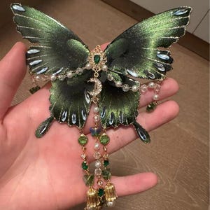 Puede incluir: Un adorno decorativo de mariposa en verde met&aacute;lico oscuro con bordes brillantes, decorado con cristales verdes y transparentes, perlas blancas y un dije de media luna dorado. Tres cadenas colgantes presentan cuentas verdes y dijes dorados en forma de campana.