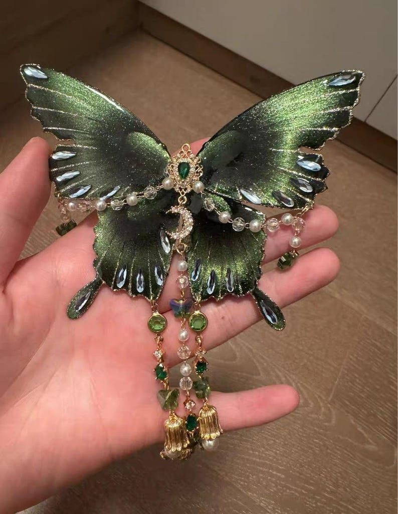 Puede incluir: Un adorno decorativo de mariposa en verde met&aacute;lico oscuro con bordes brillantes, decorado con cristales verdes y transparentes, perlas blancas y un dije de media luna dorado. Tres cadenas colgantes presentan cuentas verdes y dijes dorados en forma de campana.