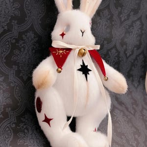 Puede incluir: Un conejo de peluche blanco con detalles de estrellas rojas. Tiene un cuello rojo y blanco con una campana dorada y una estrella negra en el pecho. Las orejas del conejo est&aacute;n adornadas con soles dorados y gemas rojas. Un art&iacute;culo decorativo.