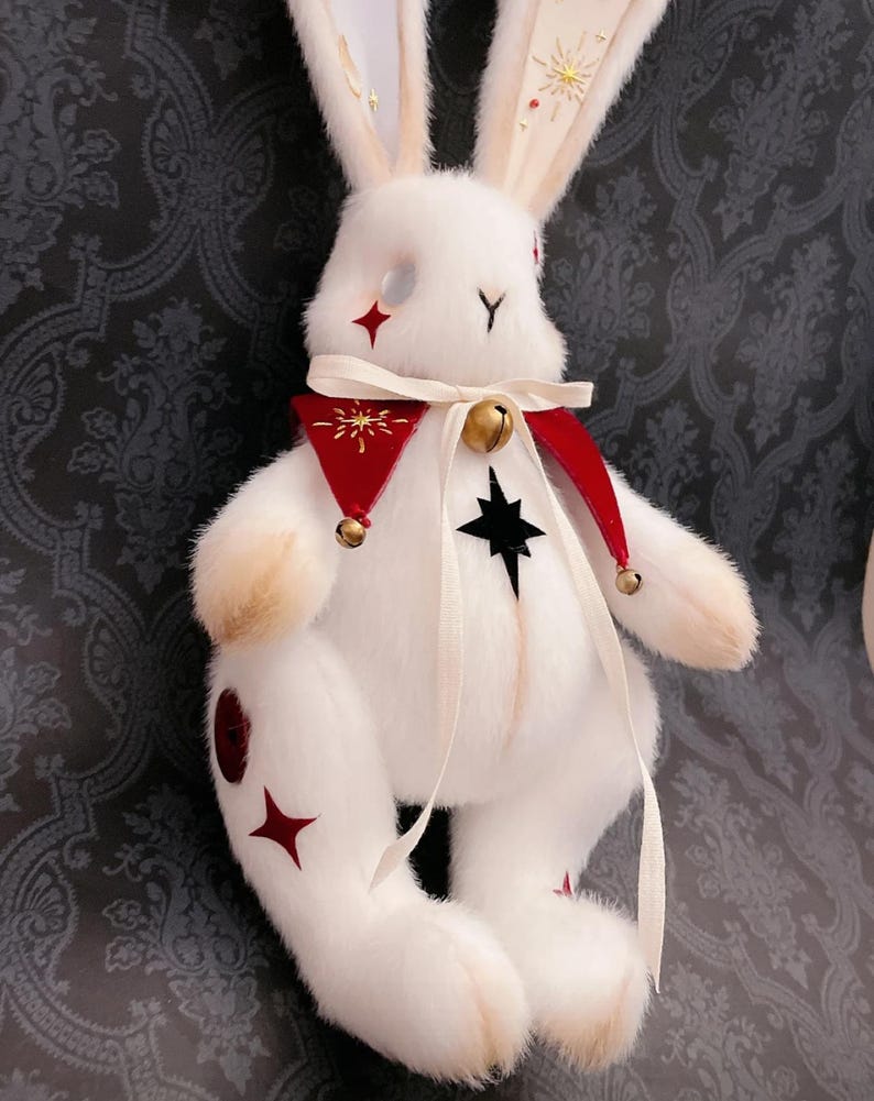 Puede incluir: Un conejo de peluche blanco con detalles de estrellas rojas. Tiene un cuello rojo y blanco con una campana dorada y una estrella negra en el pecho. Las orejas del conejo est&aacute;n adornadas con soles dorados y gemas rojas. Un art&iacute;culo decorativo.