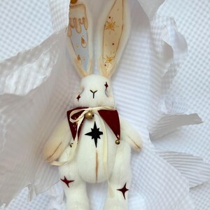 Puede incluir: Una mu&ntilde;eca de peluche de conejo blanco con orejas largas, un cuello burdeos y detalles de estrellas. Las orejas del conejo est&aacute;n decoradas con dise&ntilde;os de velas y estrellas. La mu&ntilde;eca tiene una estrella negra en el pecho y detalles de estrellas rojas en las piernas.