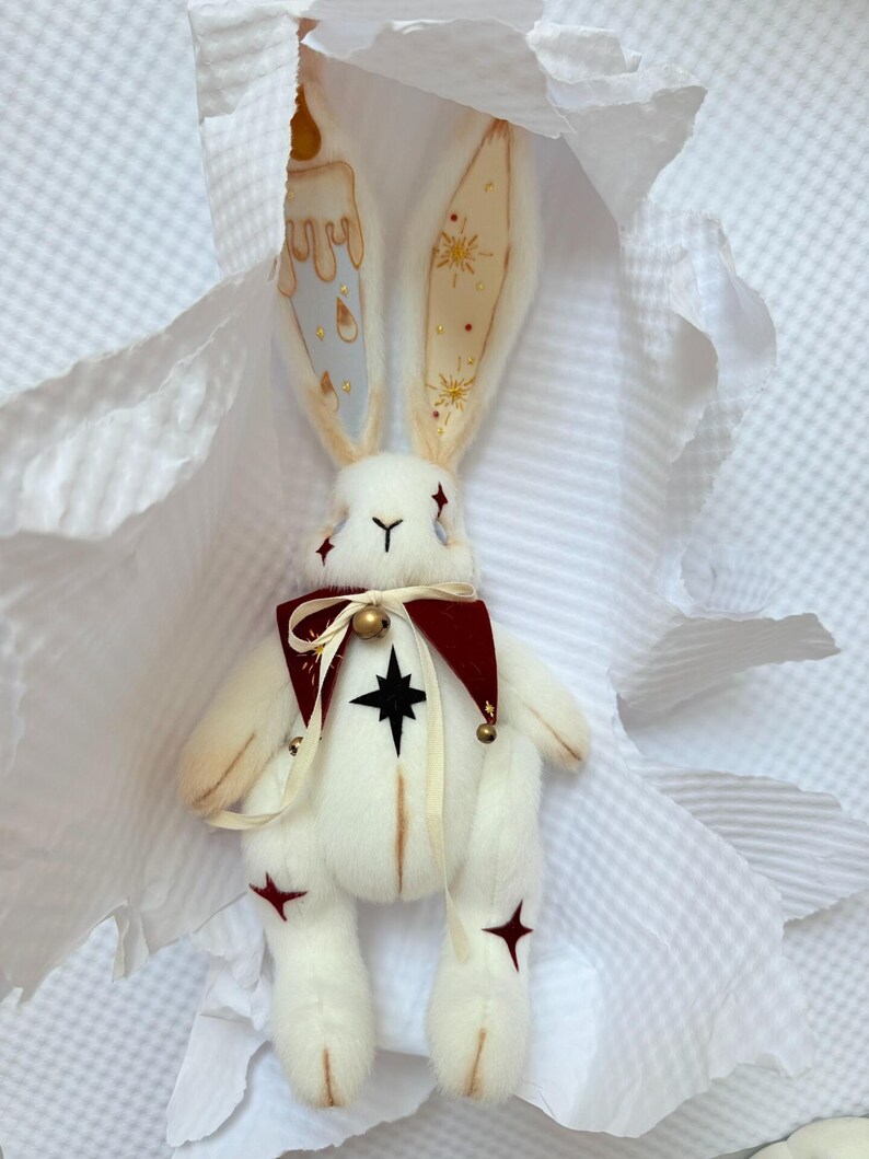 Puede incluir: Una mu&ntilde;eca de peluche de conejo blanco con orejas largas, un cuello burdeos y detalles de estrellas. Las orejas del conejo est&aacute;n decoradas con dise&ntilde;os de velas y estrellas. La mu&ntilde;eca tiene una estrella negra en el pecho y detalles de estrellas rojas en las piernas.