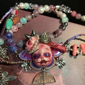 May include: A unique necklace featuring a central pendant with a doll-like face, adorned with a crown and pearl accents. The pendant is surrounded by colorful beads and charms, including a butterfly and a bow. The color palette includes pink, purple, and silver.