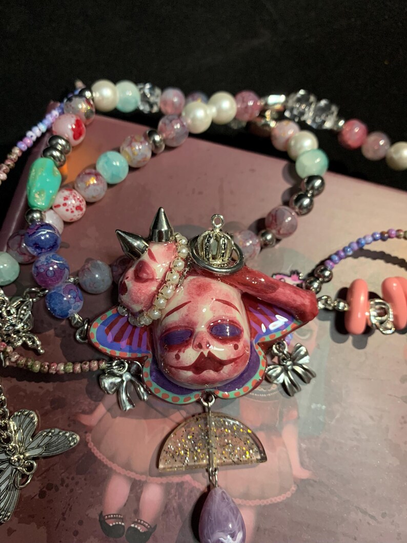 May include: A unique necklace featuring a central pendant with a doll-like face, adorned with a crown and pearl accents. The pendant is surrounded by colorful beads and charms, including a butterfly and a bow. The color palette includes pink, purple, and silver.