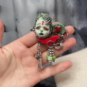 May include: A unique brooch featuring a stylized face with red eyes, green skin, and dark green lips. The brooch has a red cross and a red accent. Silver-toned metal accents and dangling charms complete the design.