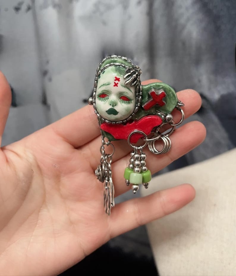 May include: A unique brooch featuring a stylized face with red eyes, green skin, and dark green lips. The brooch has a red cross and a red accent. Silver-toned metal accents and dangling charms complete the design.
