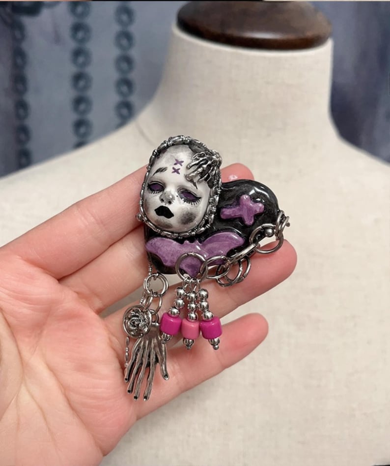 May include: A handcrafted brooch featuring a doll face with a black and silver color scheme. The brooch has a heart-shaped design with a purple cross and dangling pink beads. The brooch is held in a hand.