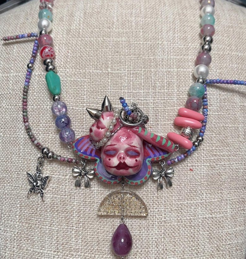 May include: A unique necklace featuring a central pendant with a pink, doll-like face and a butterfly-shaped design. The necklace incorporates various beads in shades of pink, purple, and blue, along with silver accents and a teardrop-shaped purple gem.