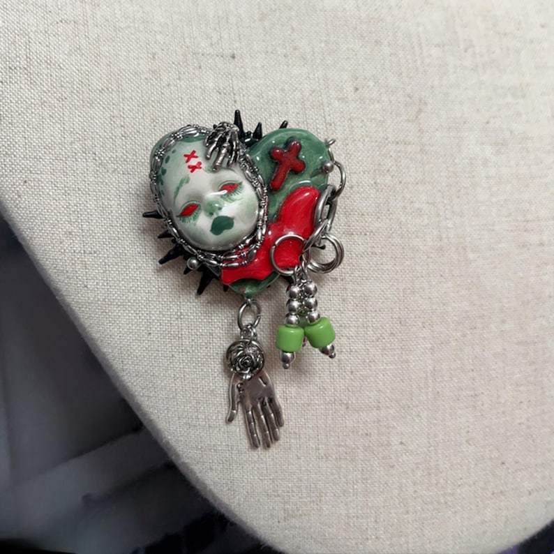 May include: A heart-shaped brooch with a doll face, red accents, and green details. The brooch features a hand charm, green beads, and a cross design. The edges are lined with spikes and connected with a chain.