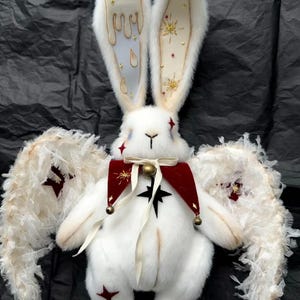 Puede incluir: Un conejo de peluche blanco con orejas largas, adornado con dise&ntilde;os de velas y estrellas. Lleva un collar rojo y negro con una campana y tiene alas de plumas. Acentos de estrellas rojas en su cuerpo.