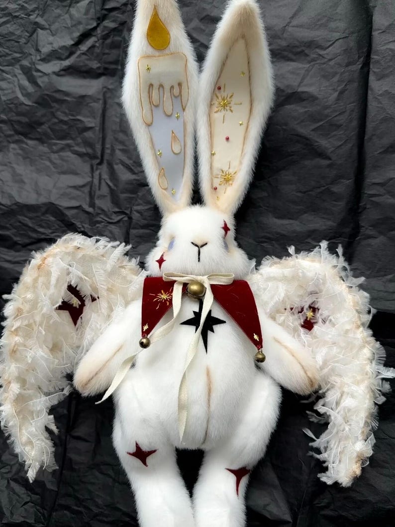 Puede incluir: Un conejo de peluche blanco con orejas largas, adornado con dise&ntilde;os de velas y estrellas. Lleva un collar rojo y negro con una campana y tiene alas de plumas. Acentos de estrellas rojas en su cuerpo.