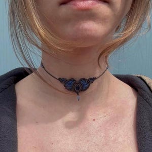 May include: A handmade macrame choker necklace. The necklace features a dark blue central design with a black stone and a small dangling crystal. The choker is made of dark brown cord and is worn around the neck.