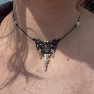 May include: A black macrame necklace with a central oval-shaped gray stone. The necklace features small silver beads and dangling silver charms. A white bone-shaped pendant hangs below the stone, adding a unique touch.